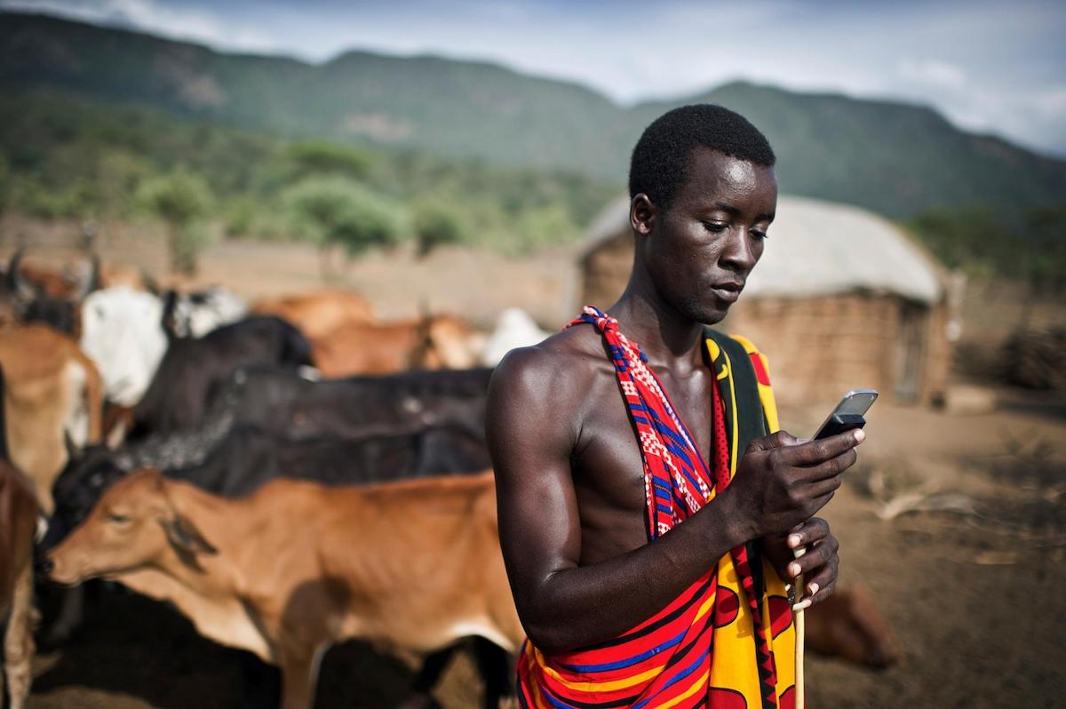 Nine year old Albert Mkatia smiles and looks down while standing in front of cattle, with his arms up and hands behind his head.