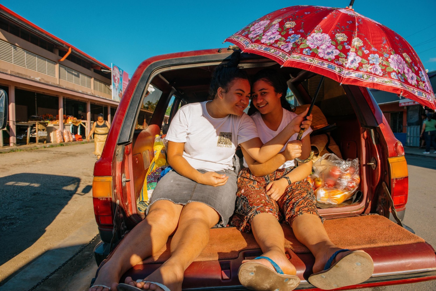 Two teen girls relaxing in the boot of a car, holding up an umbrella for shade, outside the old municipal markets in Tonga.