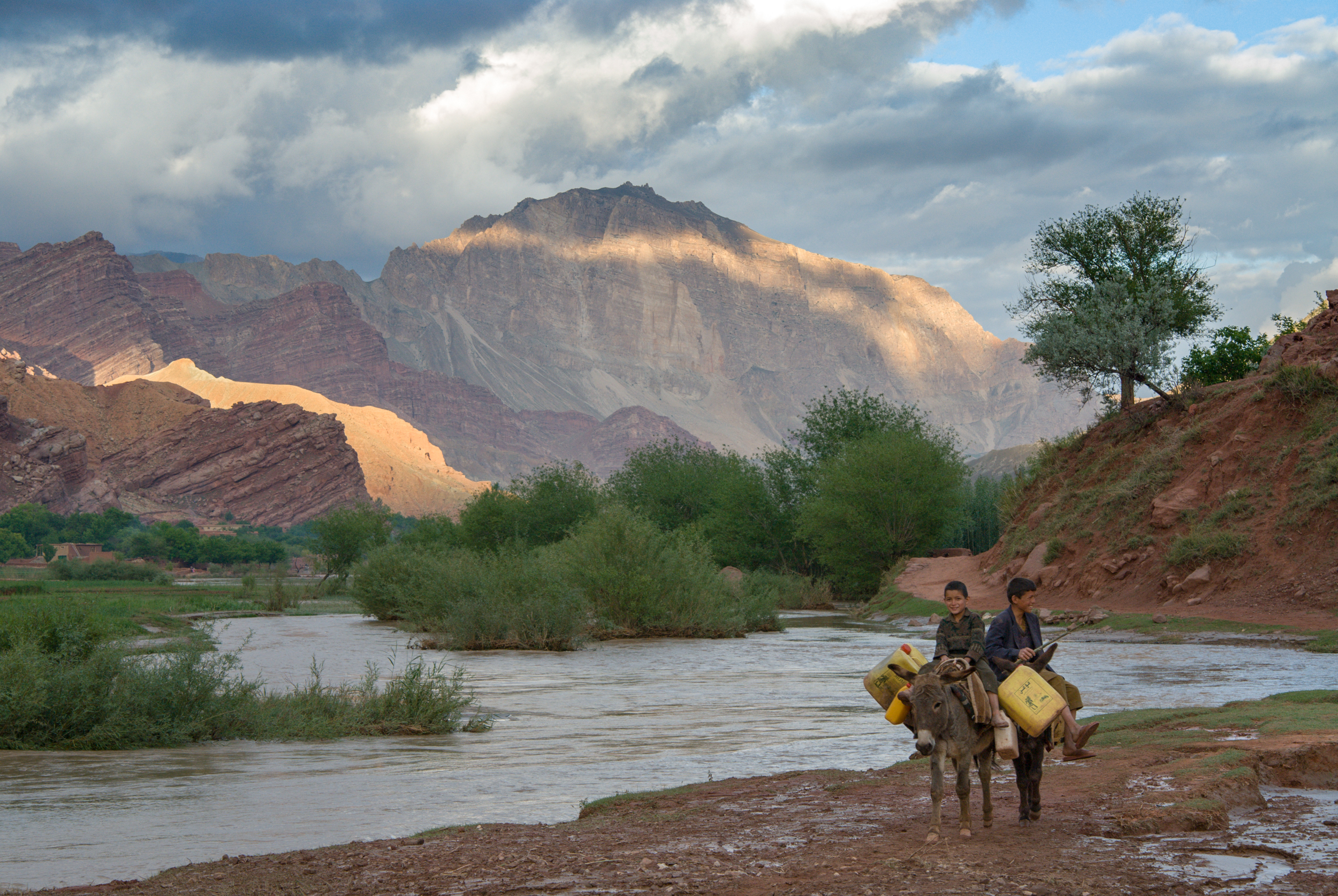 Boys on donkeys laden with water jugs walk along the edge of the Ajar river, as the surrounding red cliffs are lit by the setting sun, near Dehe Khankhala, upper Ajar Valley Bamiyan Province, Afghanistan